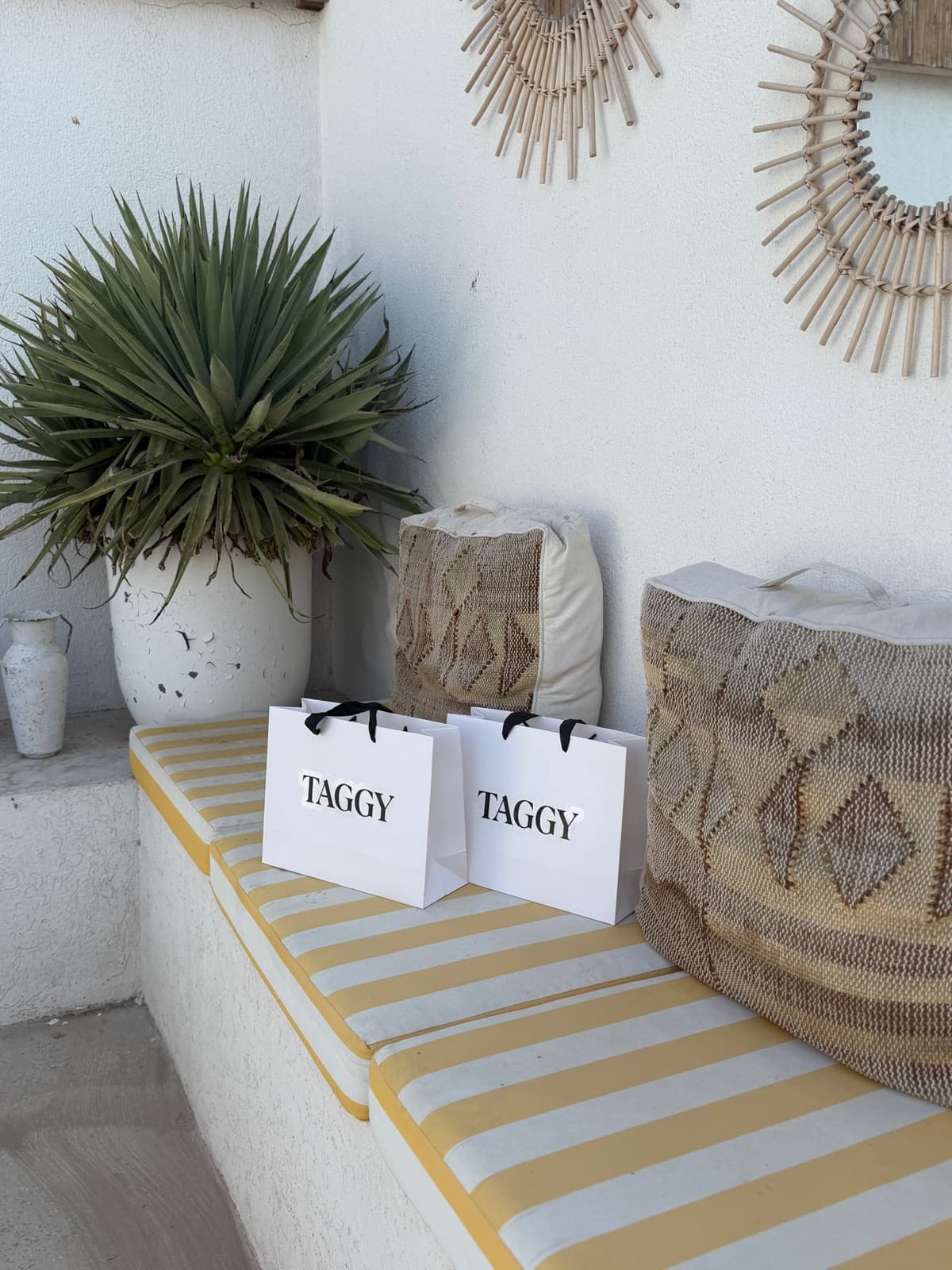 Two Taggy shopping bags on a striped bench beside woven cushions